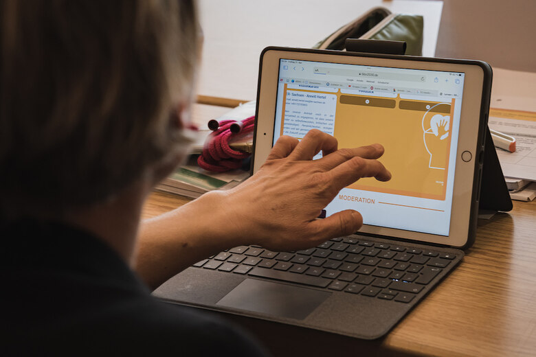 Das Bild zeigt eine Person, welche vor einem Tablet sitzt und an diesem etwas mit den Händen zeigt.