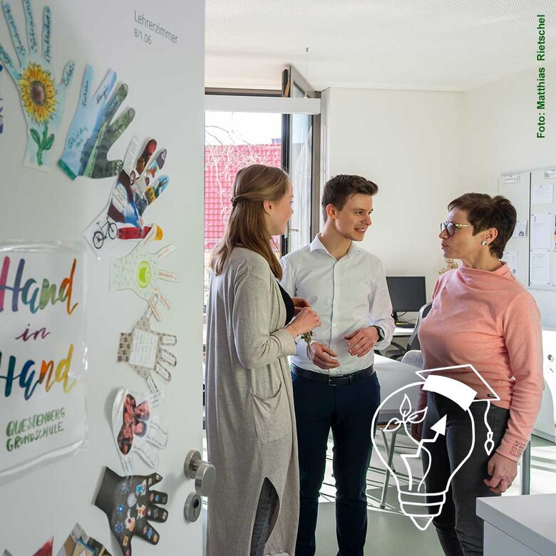 Es wird in ein Lehrerzimmer mit drei Personen geblickt. Auf der Tür sind Handabrücke zu erkennen. In der Mitte steht der Schriftzug "Hand in Hand".